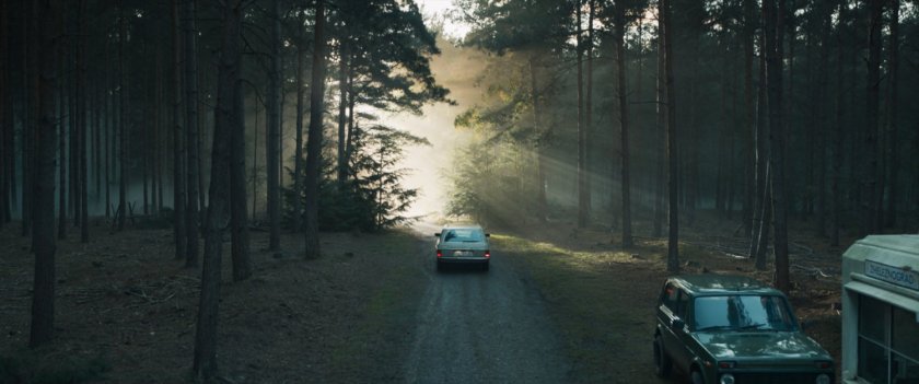 A car drives along a dirt road in a misty forest.