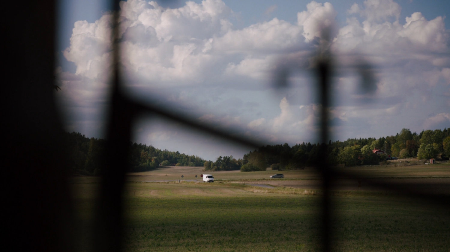 White van driving along a small country road in distance, with out of focus fence in foreground.