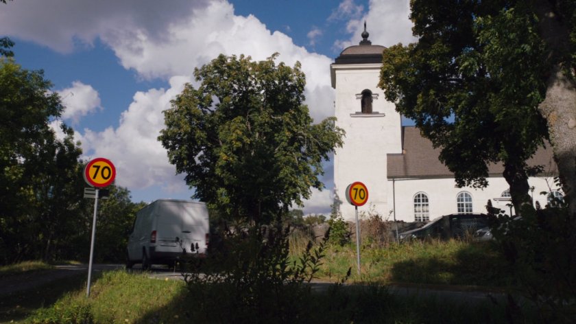 A white van drives past an old white church on a small country road.