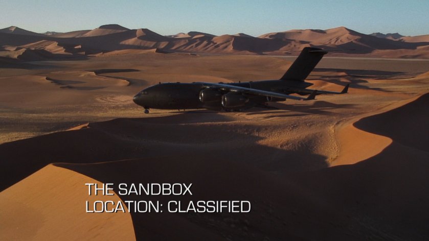 The Bus parked in the middle of sand dunes. Text: The Sandbox, Location: Classified.