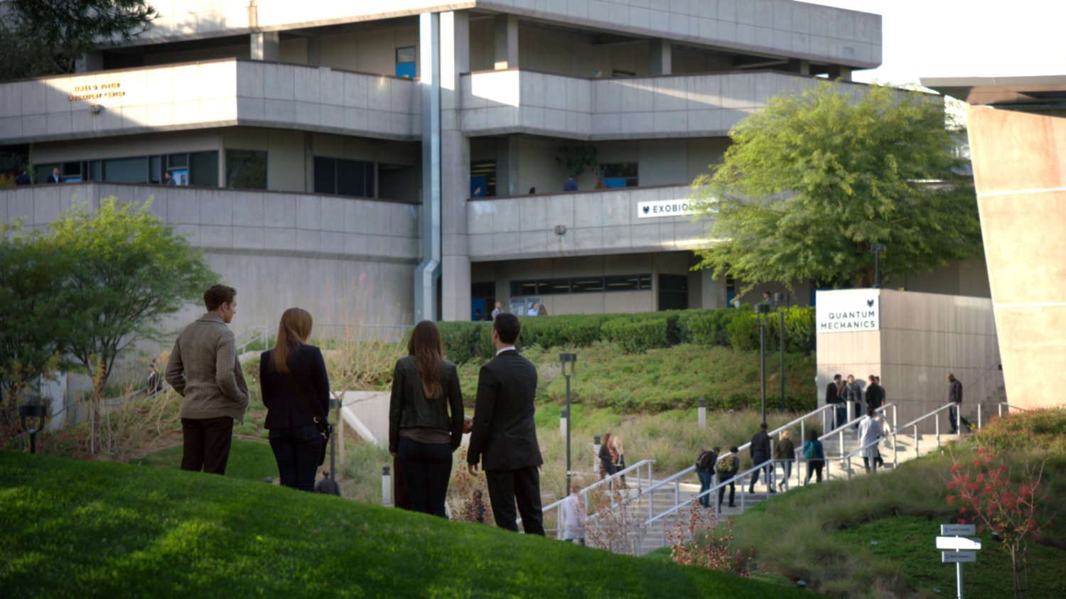 Four figures stand on a grassy hill in front of a school campus.