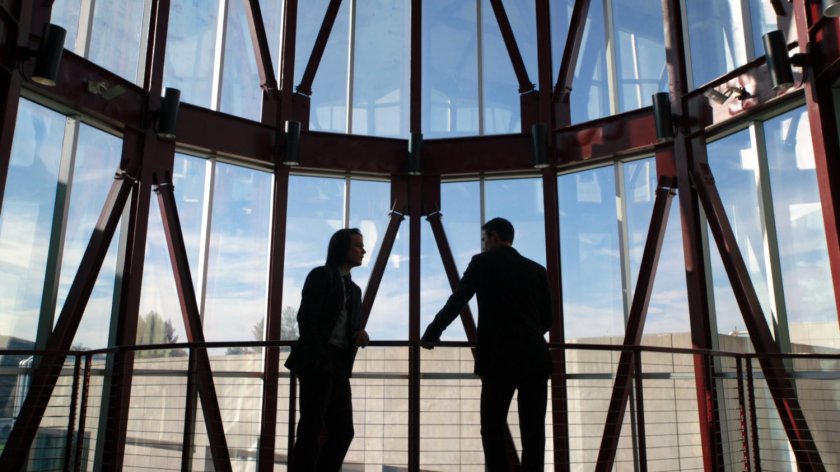 Ward and Seth talk in a small glass atrium looking onto the roof of a nearby building.