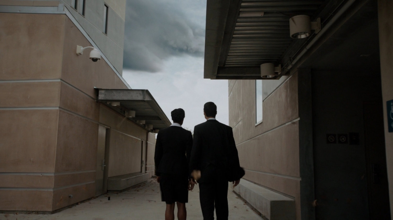 Two agents watch storm clouds from between two SHIELD Academy buildings.