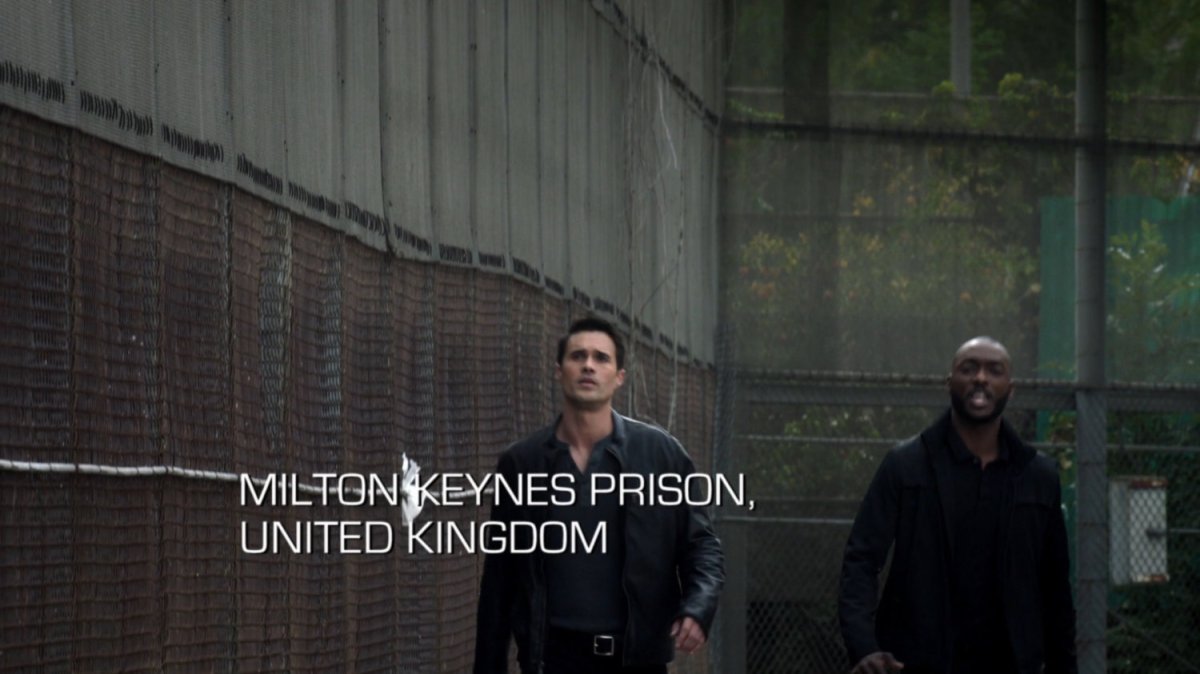 Ward and Triplett walk by a large chain link fence. Text: Milton Keynes Prison, United Kingdom.