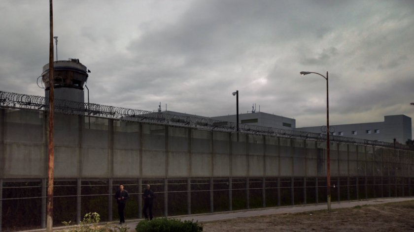 Wide shot of Ward and Triplett walking by a large chain link fence surrounding a prison.