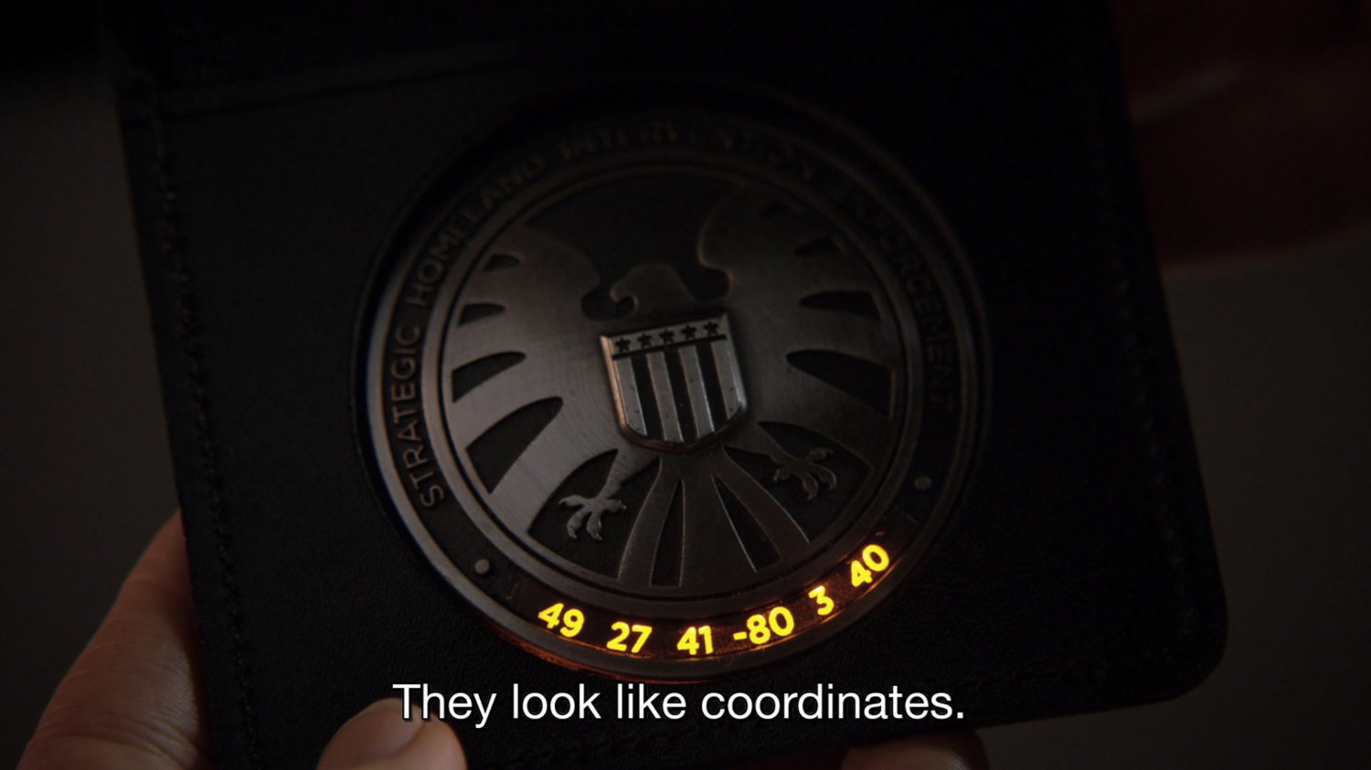 Closeup of SHIELD badge with illuminated numbers. Text: they look like coordinates.