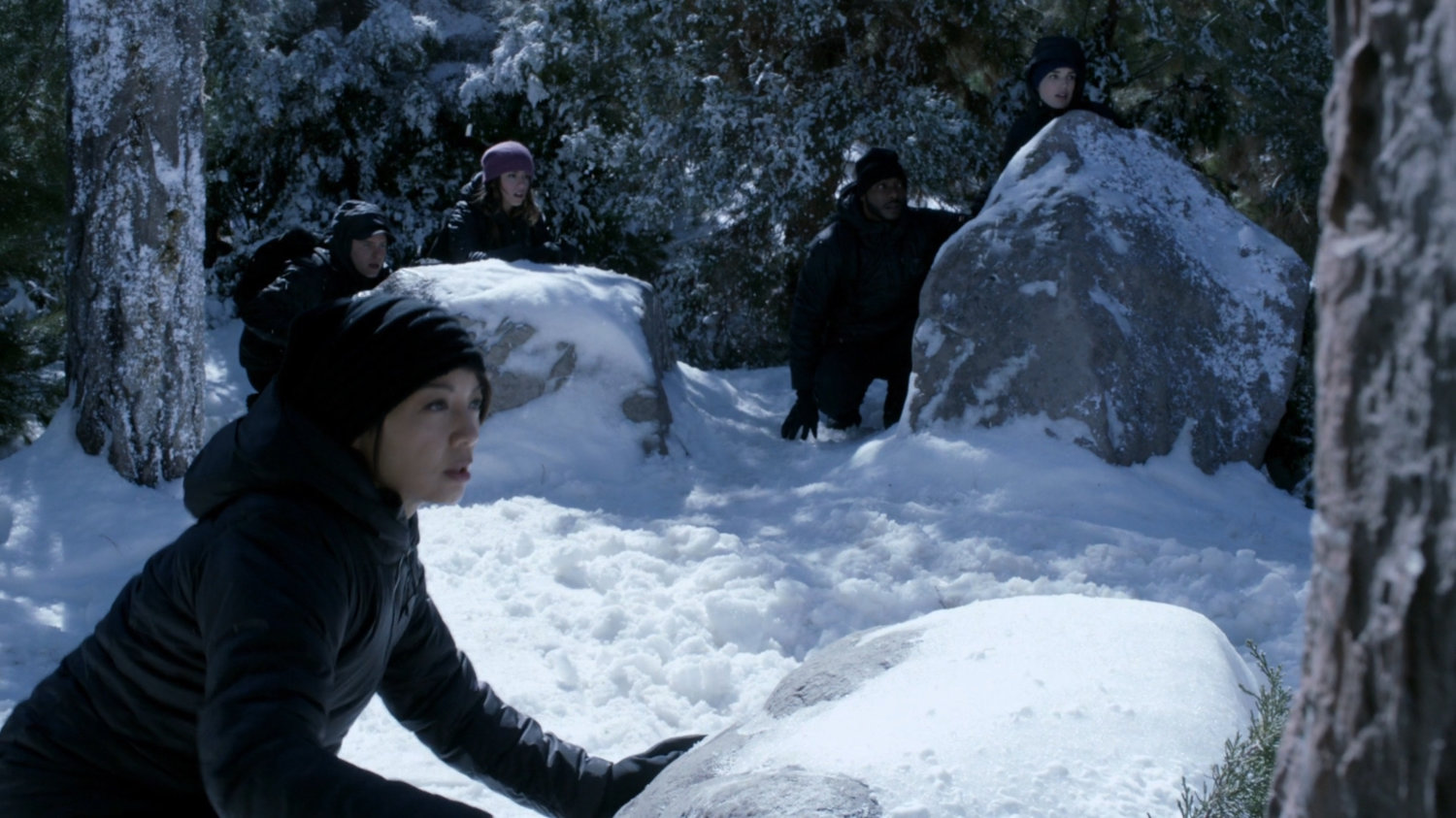 The Agents hide behind snowy rocks in the woods.