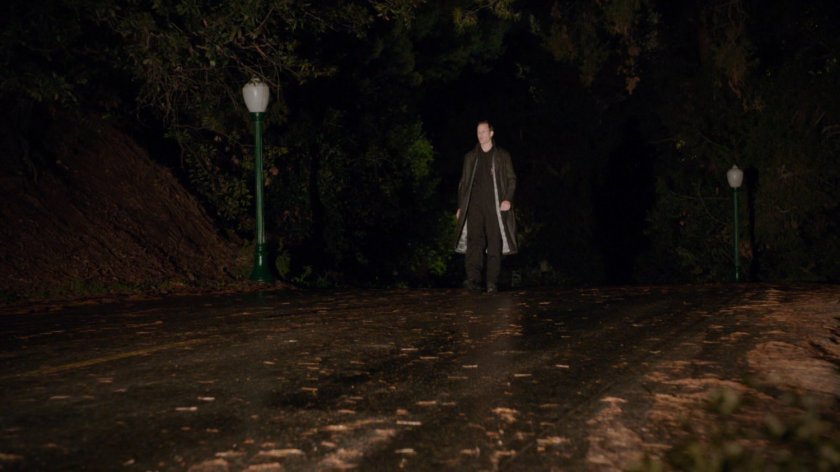 A man in black trench coat stands in the middle of a road in a Portland park.
