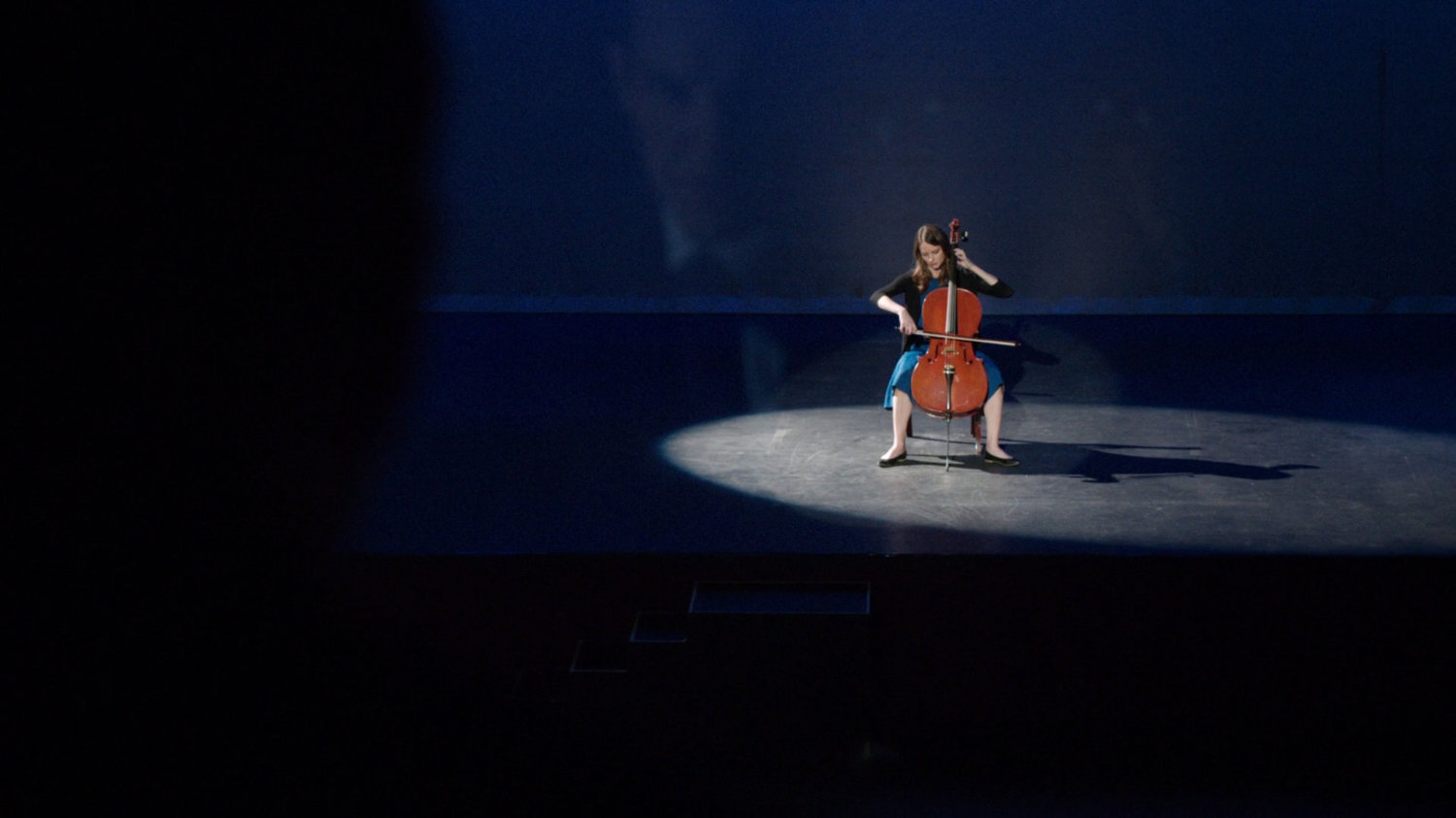 Audrey Nathan playing cello on a spotlit theater stage.