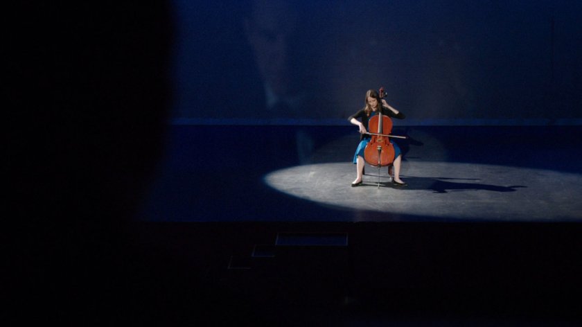 Audrey Nathan playing cello on a spotlit theater stage.