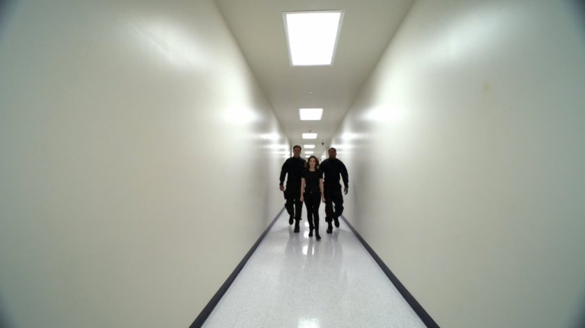 Jemma being escorted down plain white hallway with two guards.