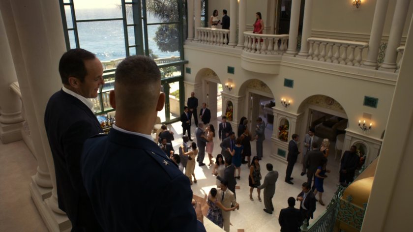 Glenn Talbot talks to Coulson on balcony in Miami mansion, while people dance below.