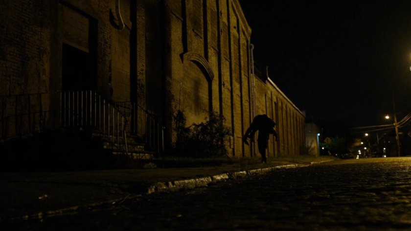 Daredevil carrying Vladimir on his shoulder along sidewalk outside abandoned warehouse.