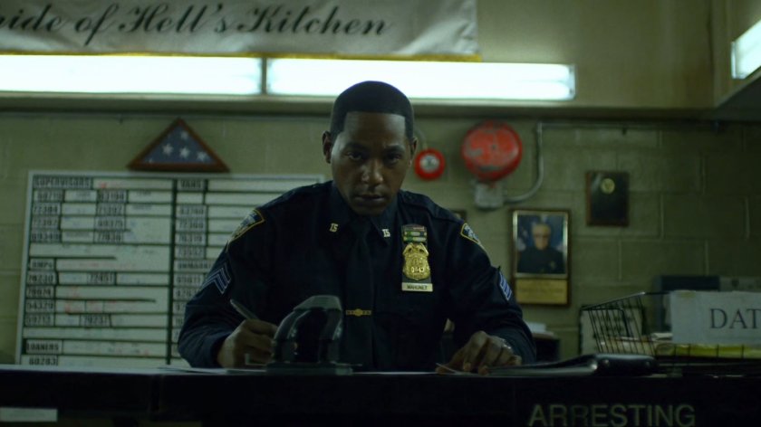 Brett Mahoney at 15th Precinct front desk. A picture of Stan Lee is in the background.