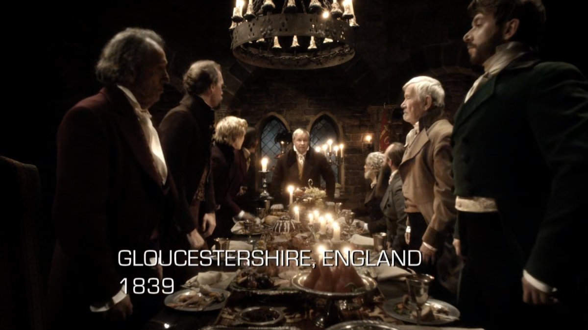 A number of men stand around a fancy table in an old castle. Text: Gloucestershire, England, 1839.