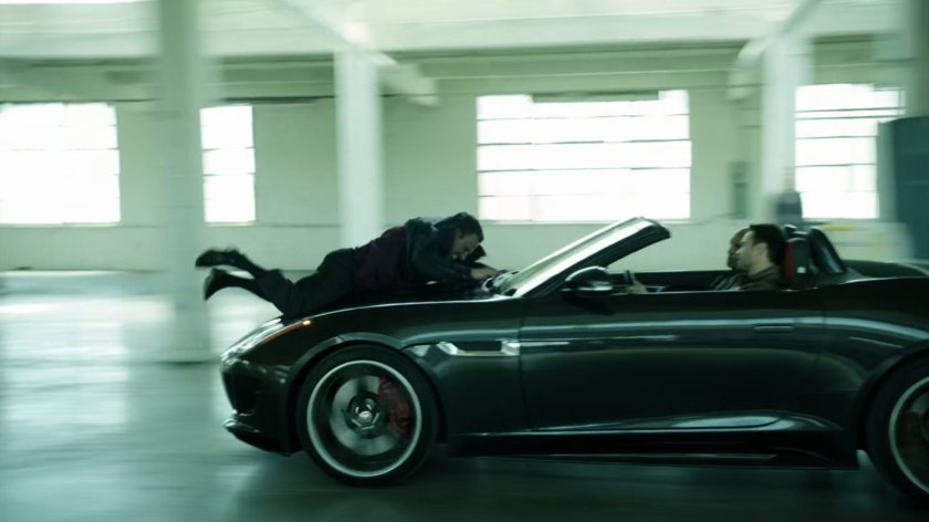A sporty car with man on the hood, drives through a warehouse.