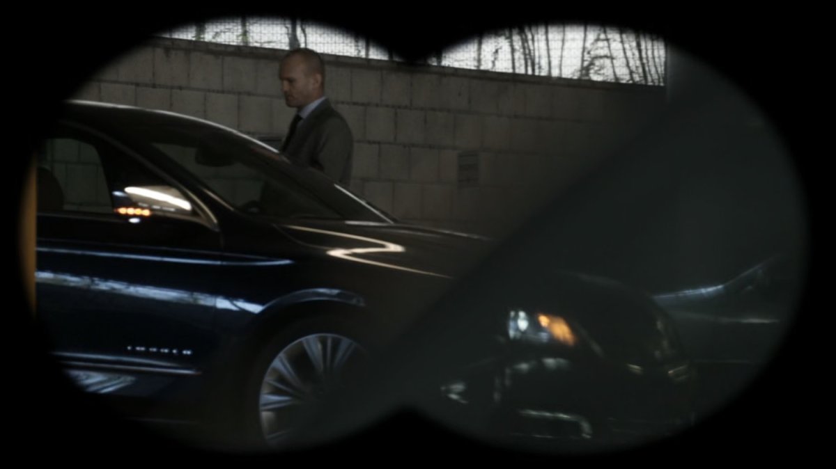 Banks getting into his car in a parking garage, as seen through binoculars.