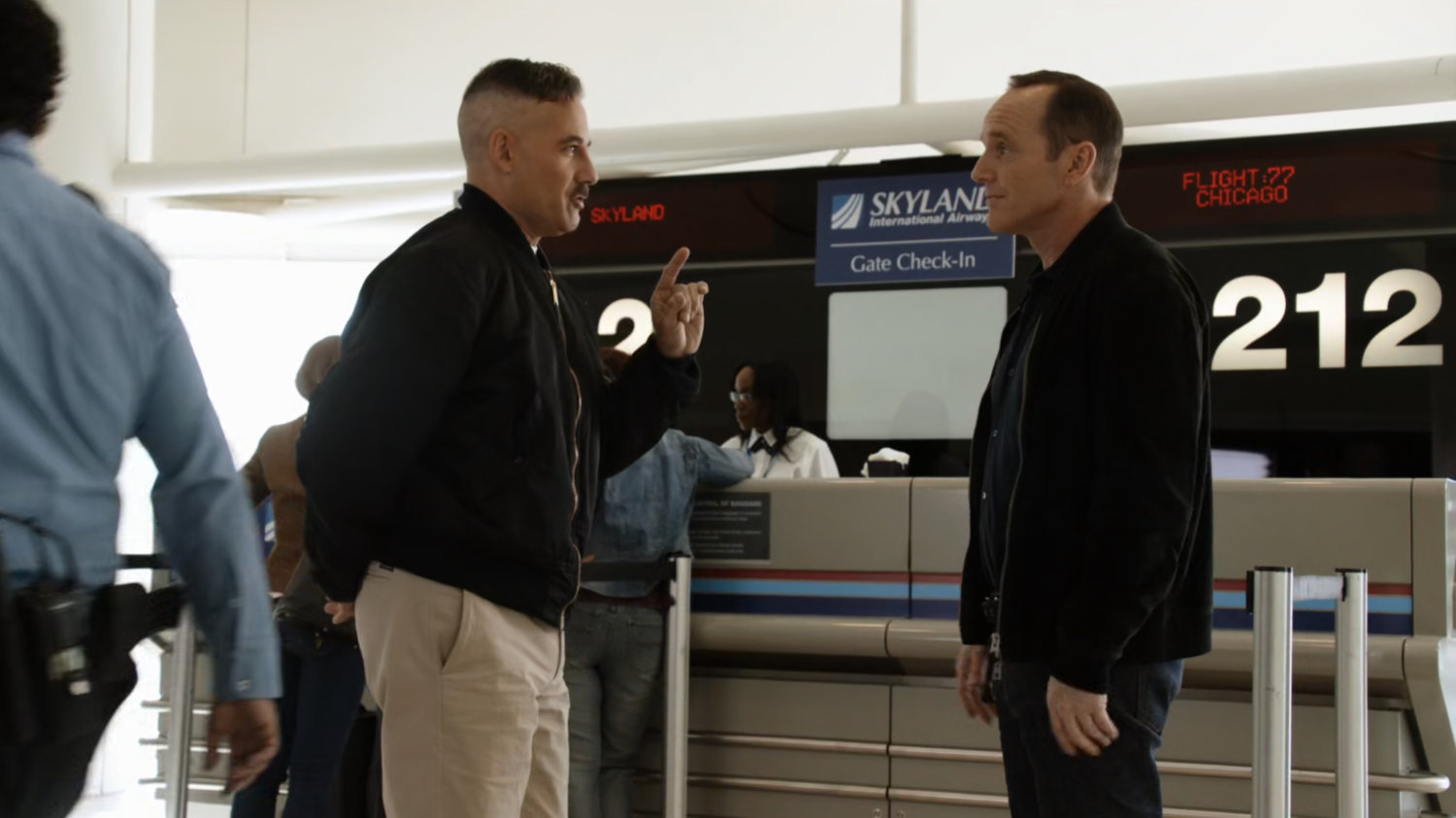 Coulson and Talbot talk in front of ticketing counter at an airport.