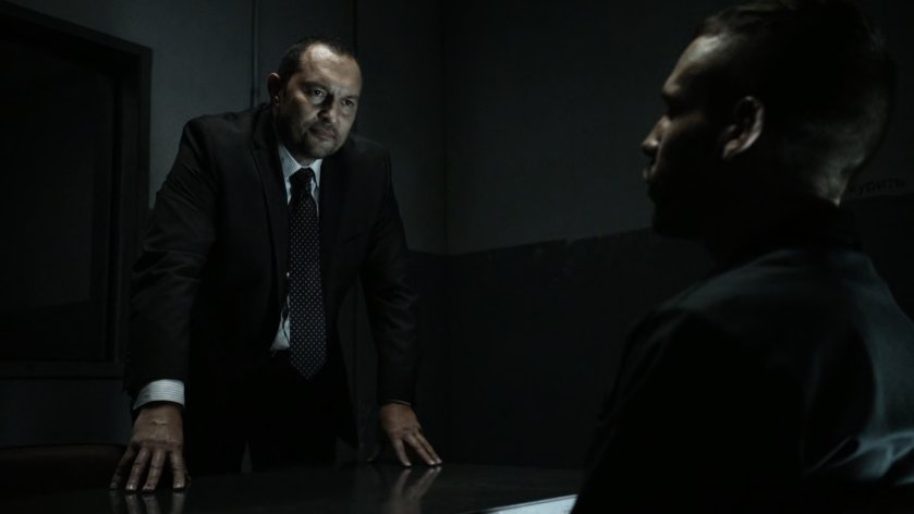 Hunter being questioned by Interpol agent in small interrogation room.