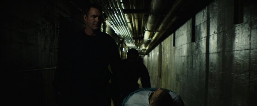 Men moving a gurney in an underground hallway with pipes overhead.