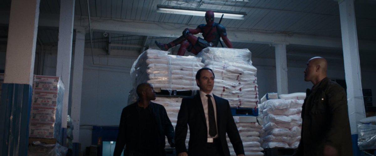 Deadpool sitting on sacks of rice above men in suits.