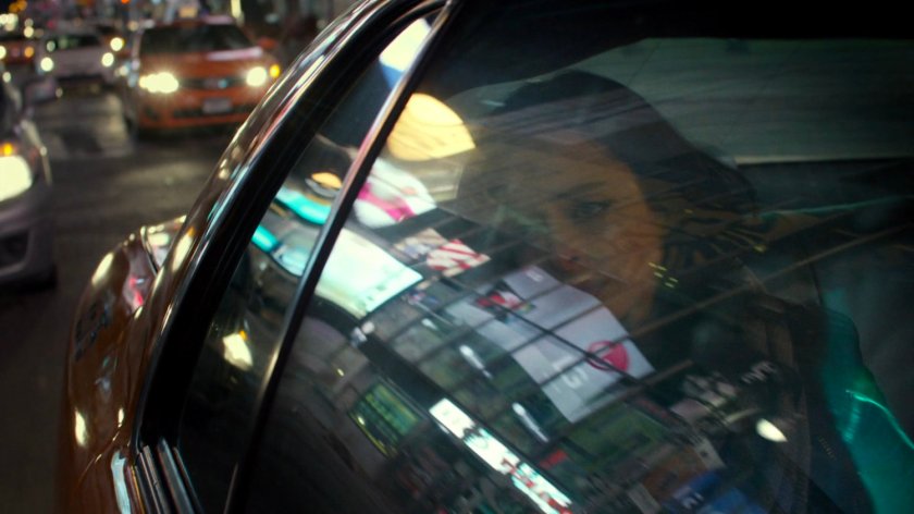 Jessica in the back seat of a taxi at night with reflections of stores in window.