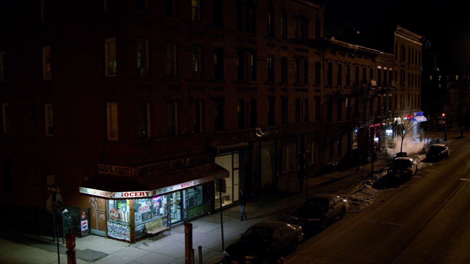 Jessica walking along a sidewalk towards corner store at night.