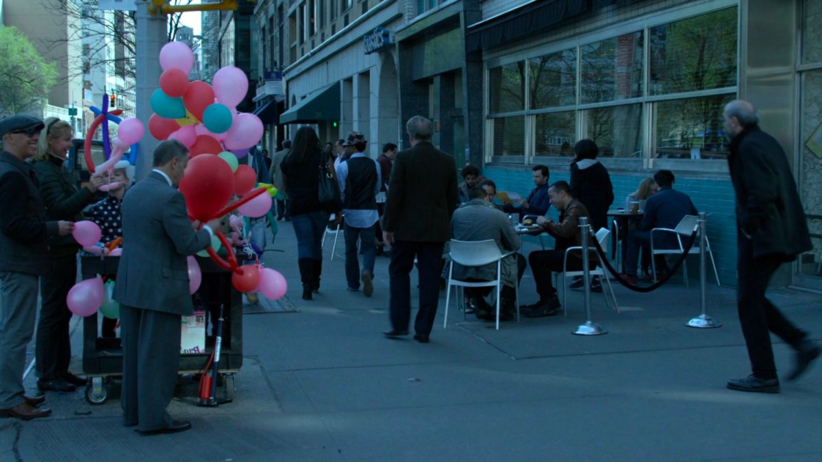 Balloon vendor and pedestrians outside corner coffee shop.