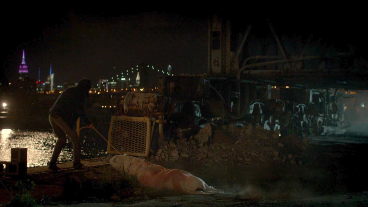Malcolm dragging a body towards a dock at night.