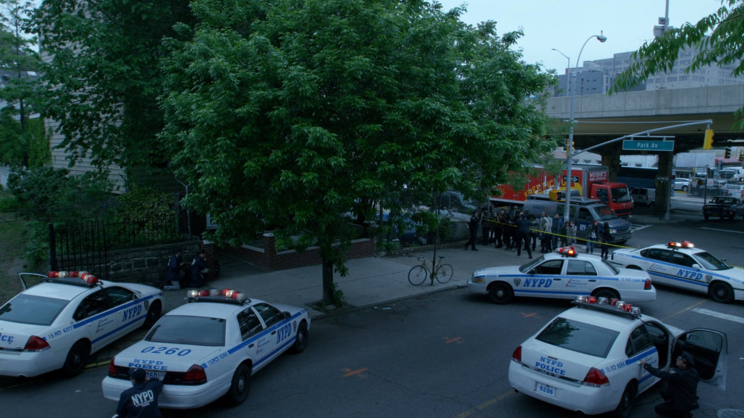 Police cars surround corner house as pedestrians look on.