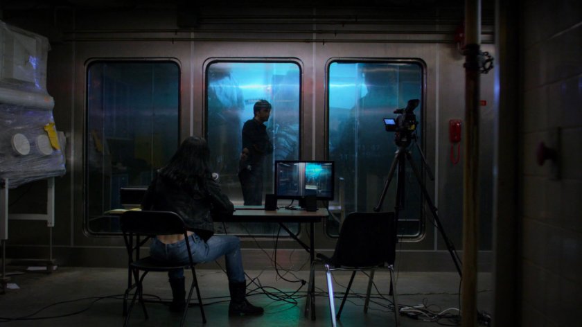 Jessica at a table looking into hermetically sealed room with Kilgrave at the CDC Facility.