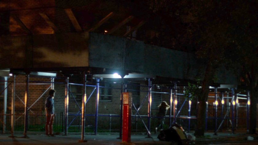 Robyn walking away from Malcolm on sidewalk under construction scaffolding at night.