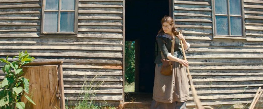 Agatha sweeps off porch outside wooden cabin, 1756.