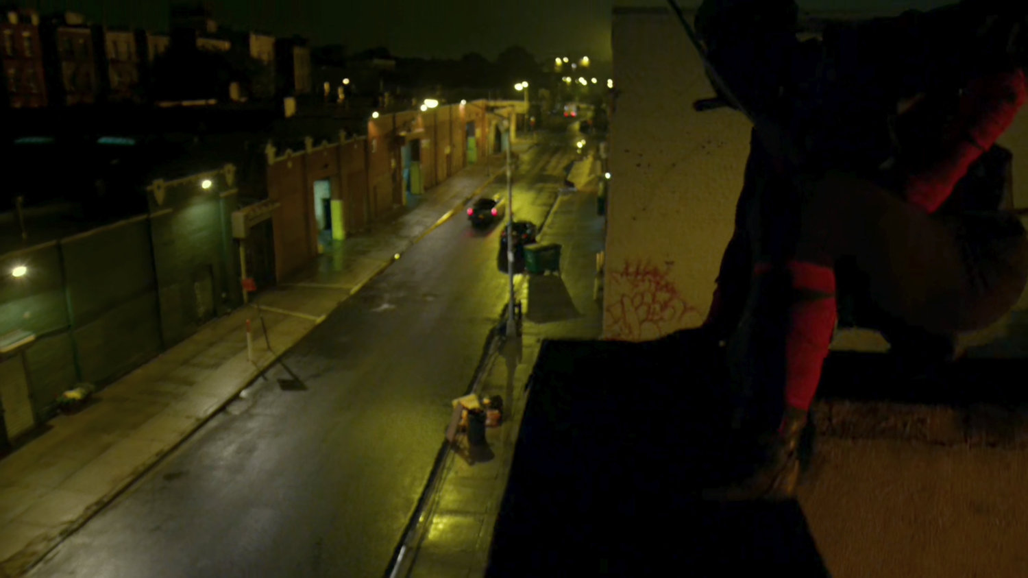 A Hand ninja atop a roof looks at Stick's car driving down a narrow street at night.