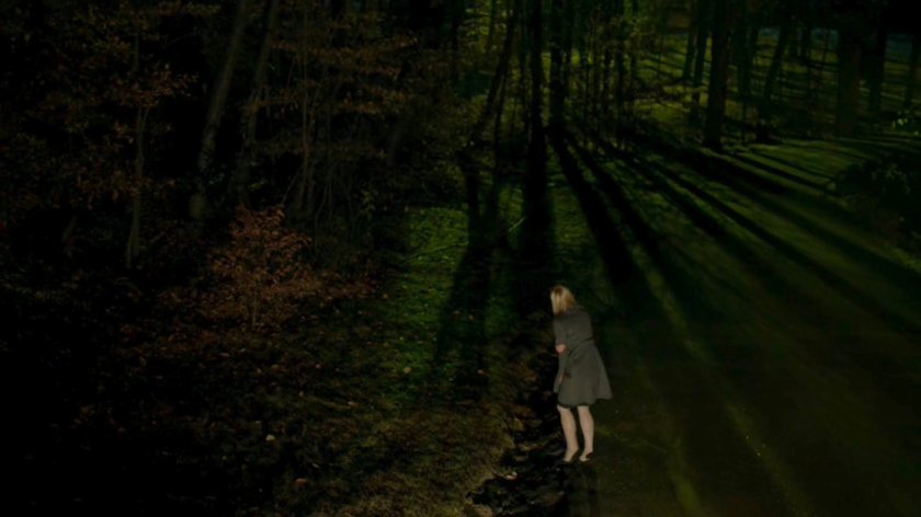 Karen entering woods from a dark, wooded road.