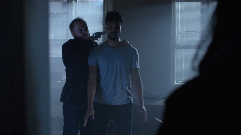 John Raymond holds Malcolm at gunpoint.