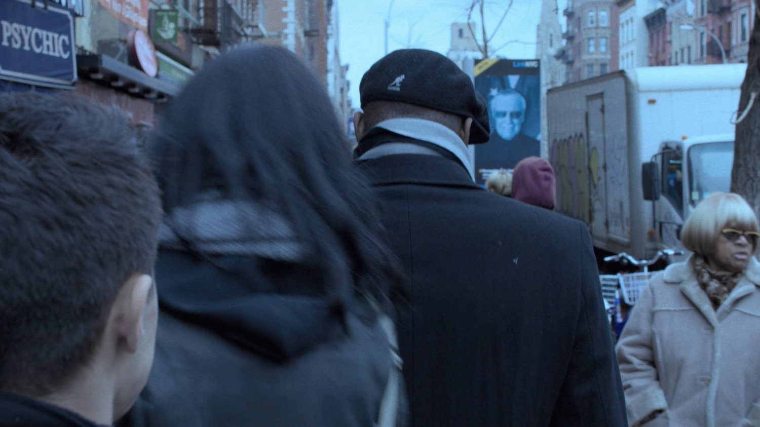 Jessica walking along a sidewalk. A cameo poster of Stan Lee is nearby.