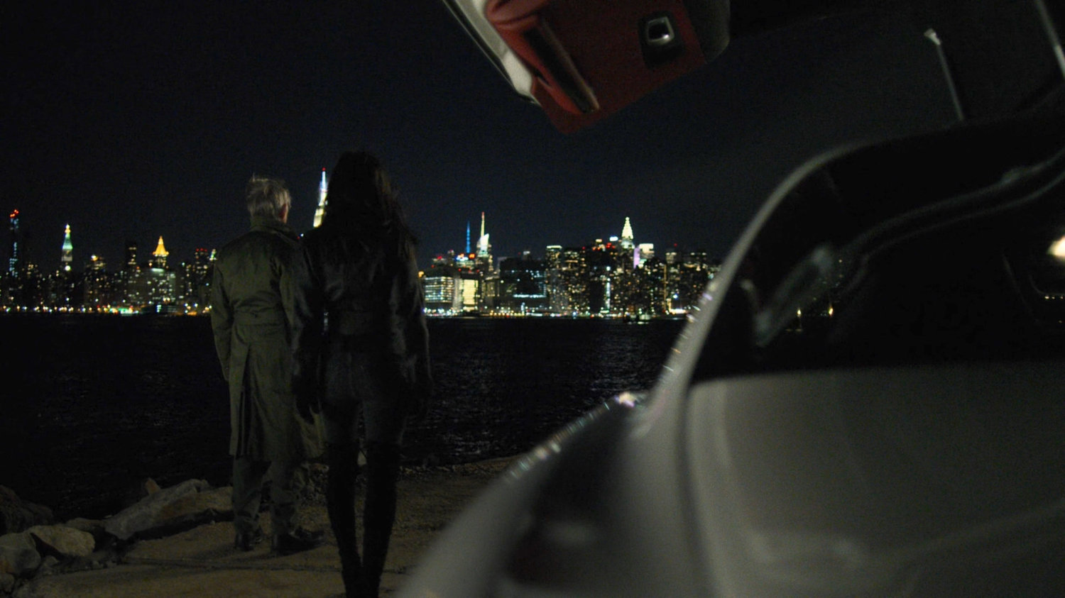 Stick and Elektra standing next to white sports car at night. A view of midtown Manhattan at night in the background.