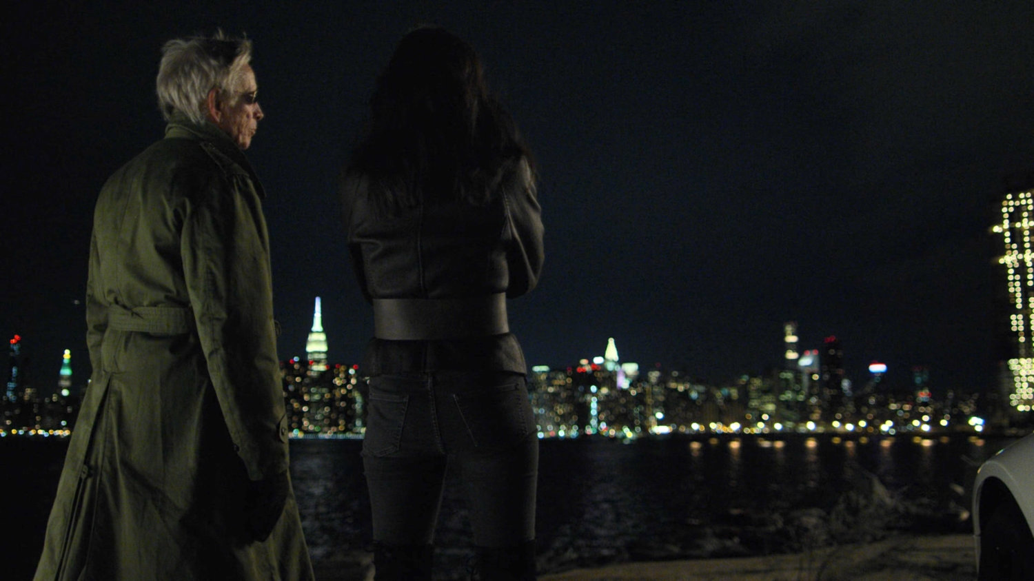 Stick and Elektra standing next to white sports car at night. A view of midtown Manhattan at night in the background.