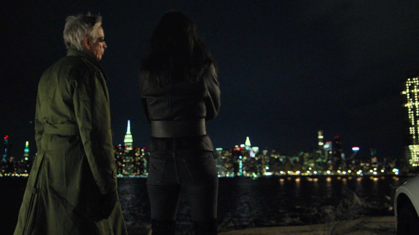 Stick and Elektra standing next to white sports car at night. A view of midtown Manhattan at night in the background.