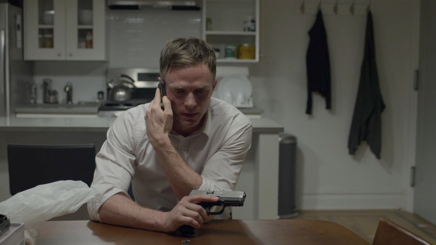 Dex with a gun, on the phone at his kitchen table.