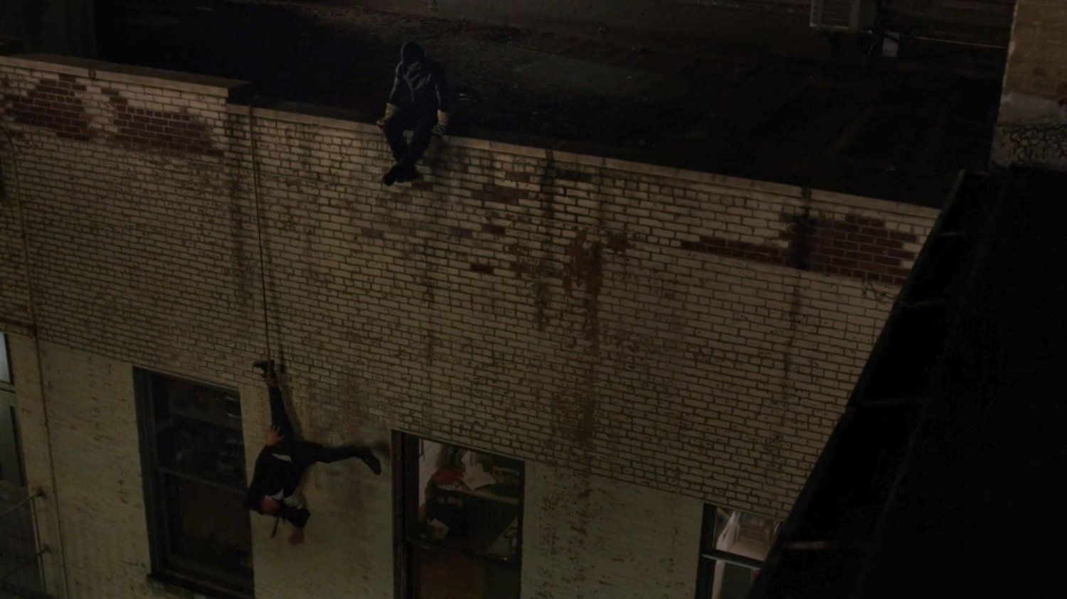 Felix Manning dangling off a dark rooftop while Matt sits on ledge above.