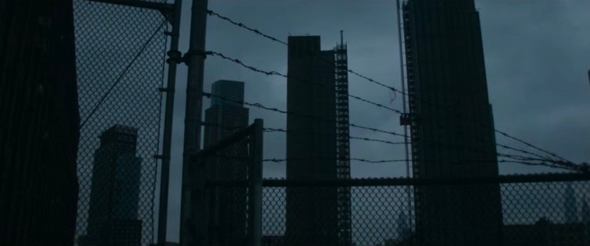 Interstitial: Tall buildings against a grey sky with barbed wire in front.