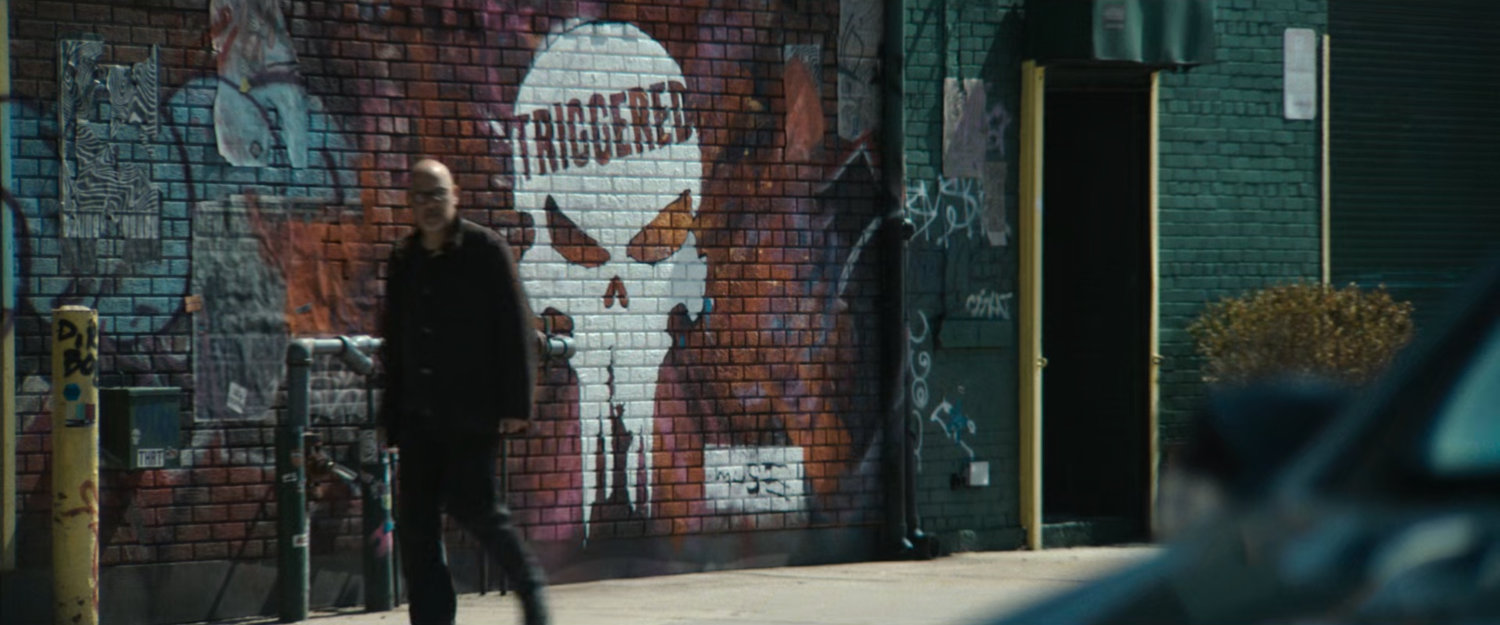 Cheery walks past a mural of the Punisher skull with word 'triggered.'