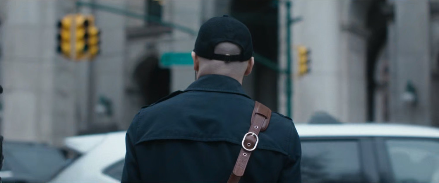 Interstitial: Man with hat and satchel standing by cars.