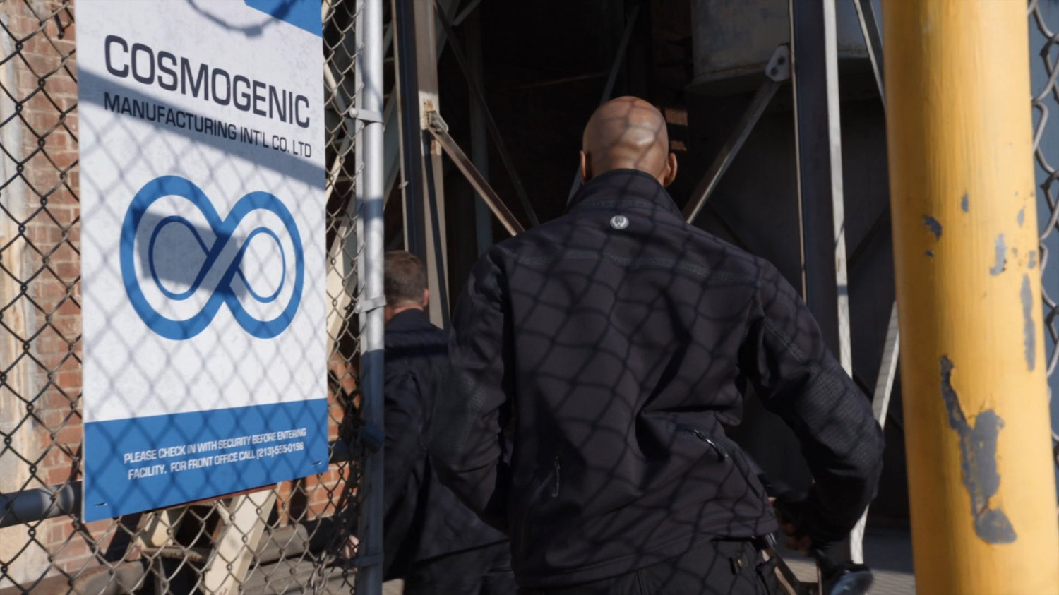 Mack and Coulson enter gate outside Cosmogenic Manufacturing.