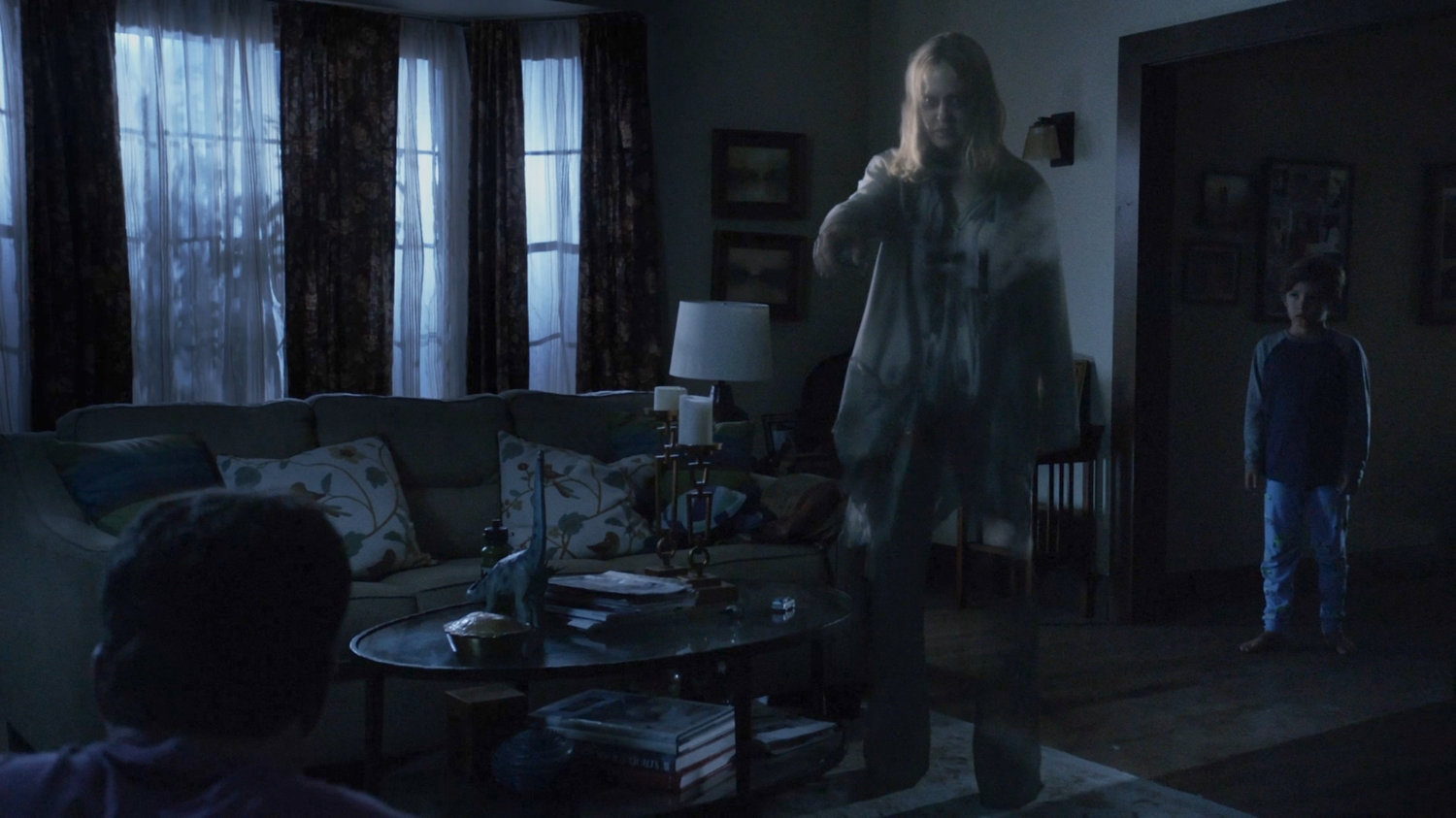 Ghost of Lucy Bauer standing in the living room pointing at the father as the young boy watches.
