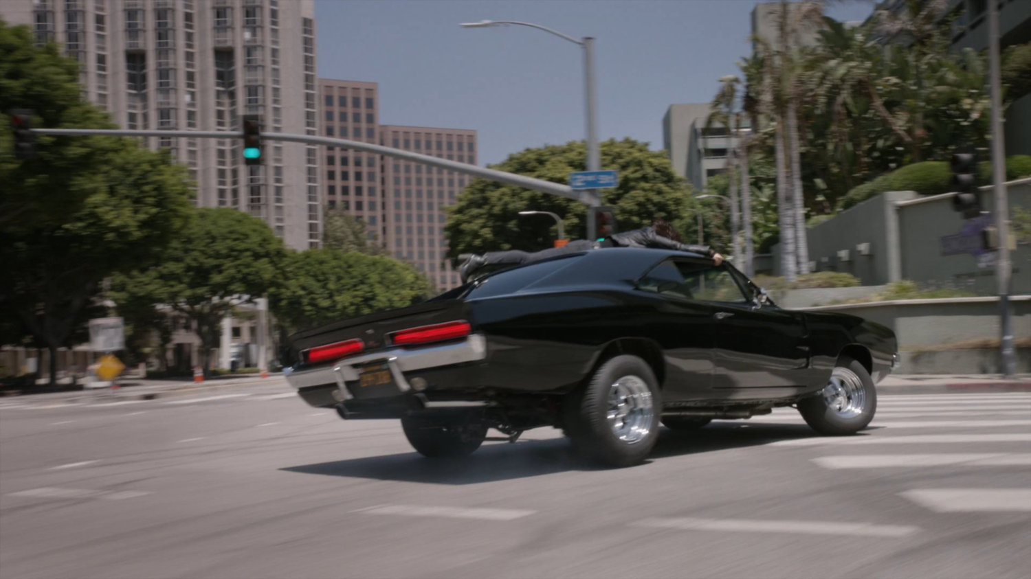 Daisy holding onto roof of Robbie's black Charger as it turns corner.