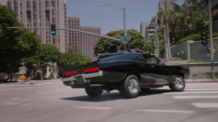Daisy holding onto roof of Robbie's black Charger as it turns corner.