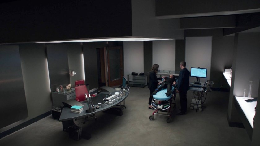 Simmons and Radcliffe stand over a gurney with Agent May in Radcliffe's house.